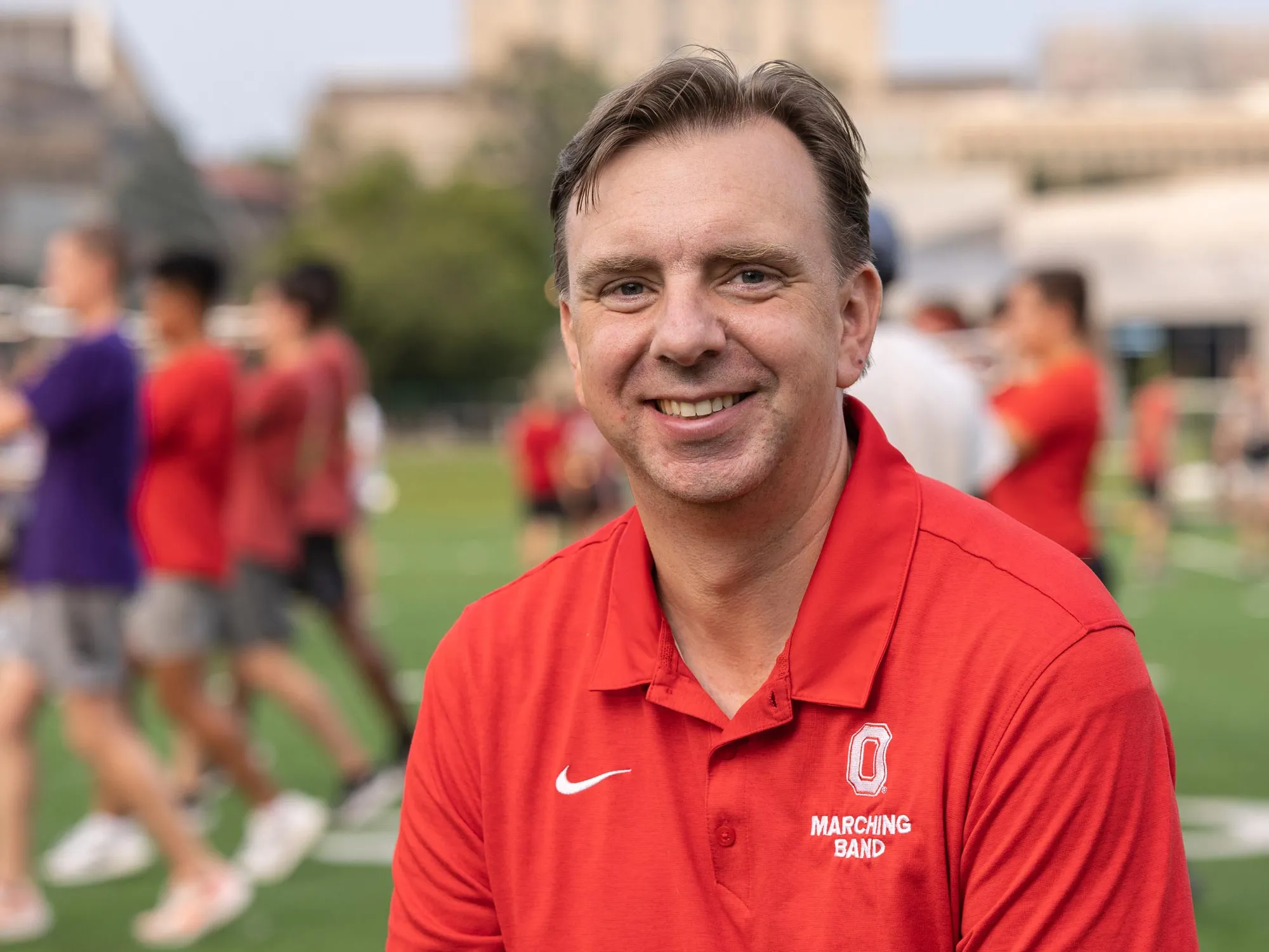 As members of the band practices behind him in their street clothes, marching band director Chris Hoch sits on a stool on the practice field. His hair’s a bit messy, his clothes are casual, and his expression is happy. 