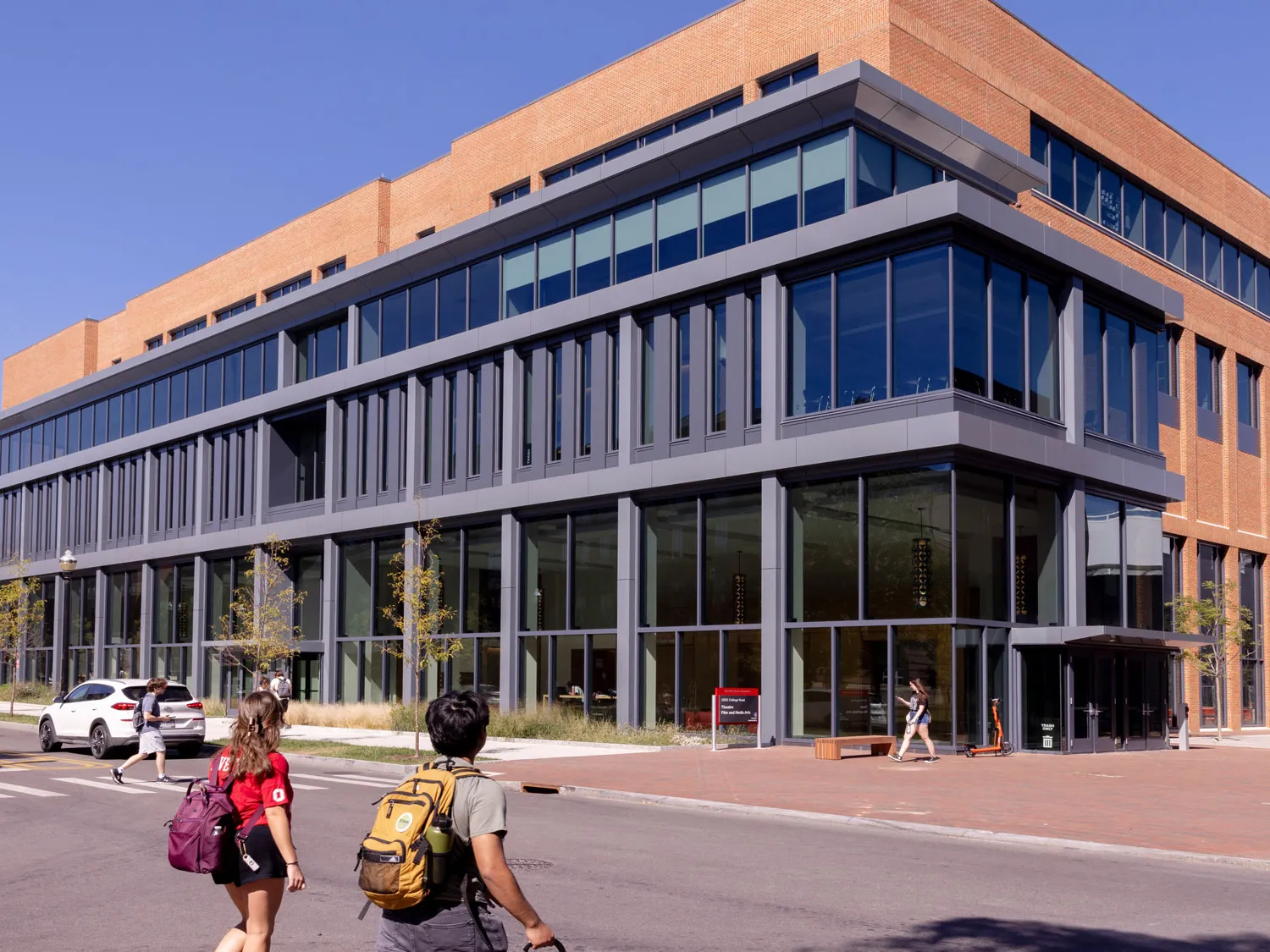 A building that is much longer than it is tall has a facade with two levels of windows and warm colored brick. It looks modern and ready for great things to come from within. Two students cross the street toward the building.
