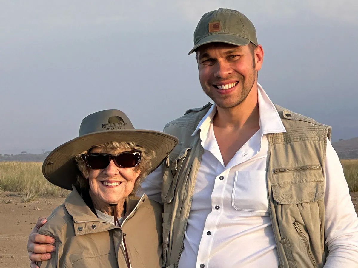 A white man stands with his arm wrapped around his much-shorter grandma. They wear khaki travelers’ clothes, hats and big smiles.