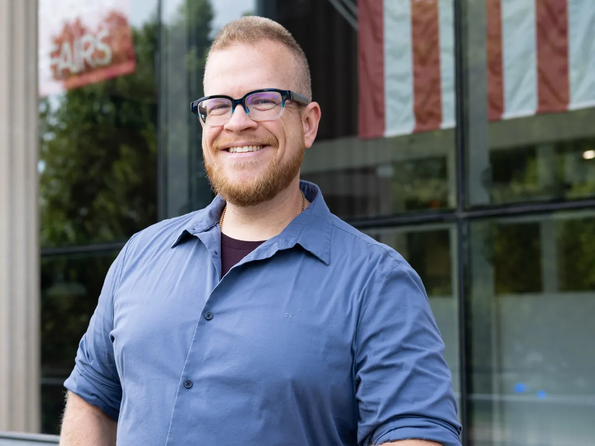 David Hibler, a broad-shouldered white man with close-cropped hair and beard and thick-frame glasses, leans on a railing outside Ohio State’s John Glenn College of Public Affairs. He has a friendly smile, the kind that crinkles his eyes, and in the background, light shines off the building’s windows. An American flag is visible depending on the reflection.