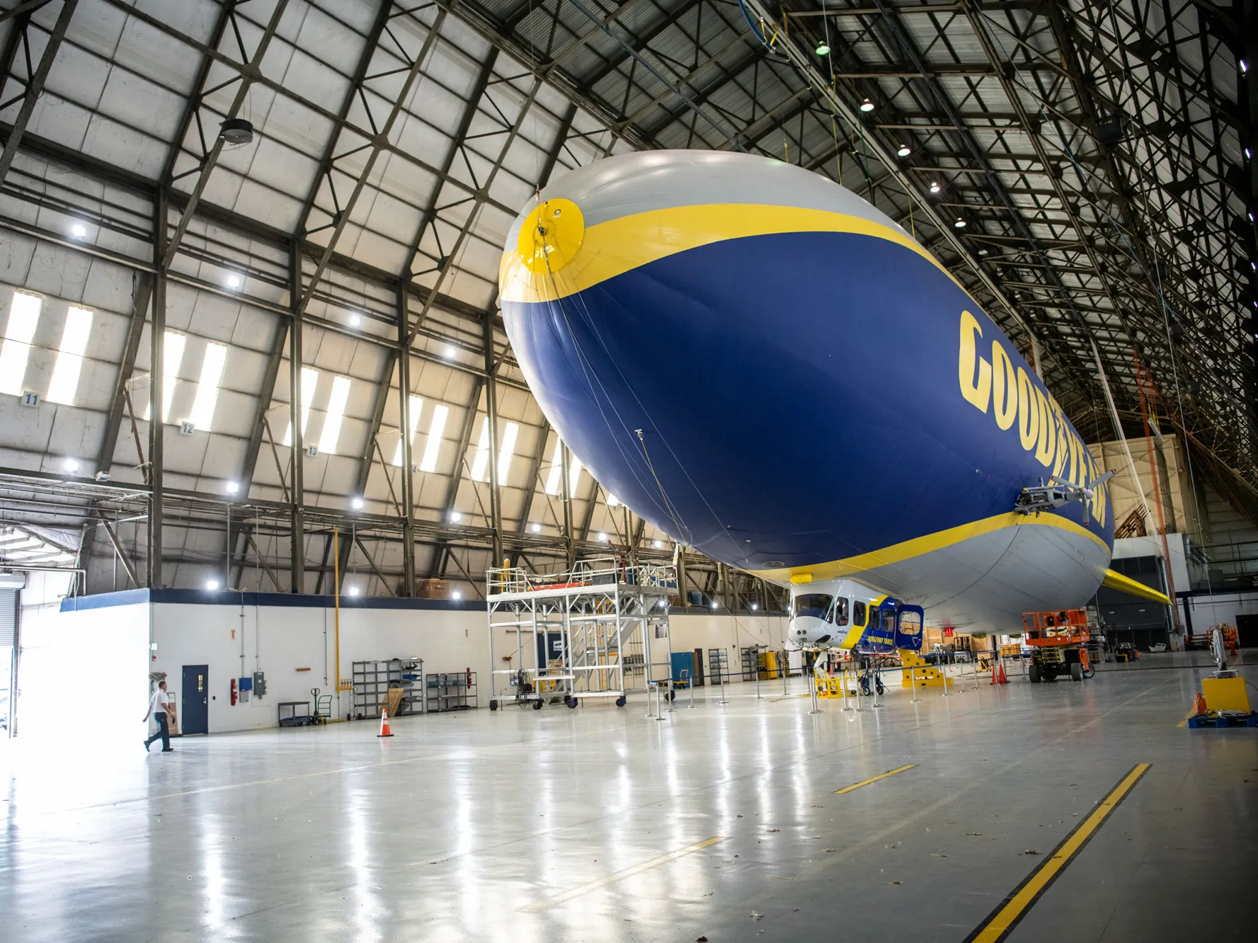 In a hangar with metal scaffolding supporting the 6-sided roof, a Goodyear blimp floats, looking a bit like a stamen inside a tulip. The floor is so clean, lights shine off it. 