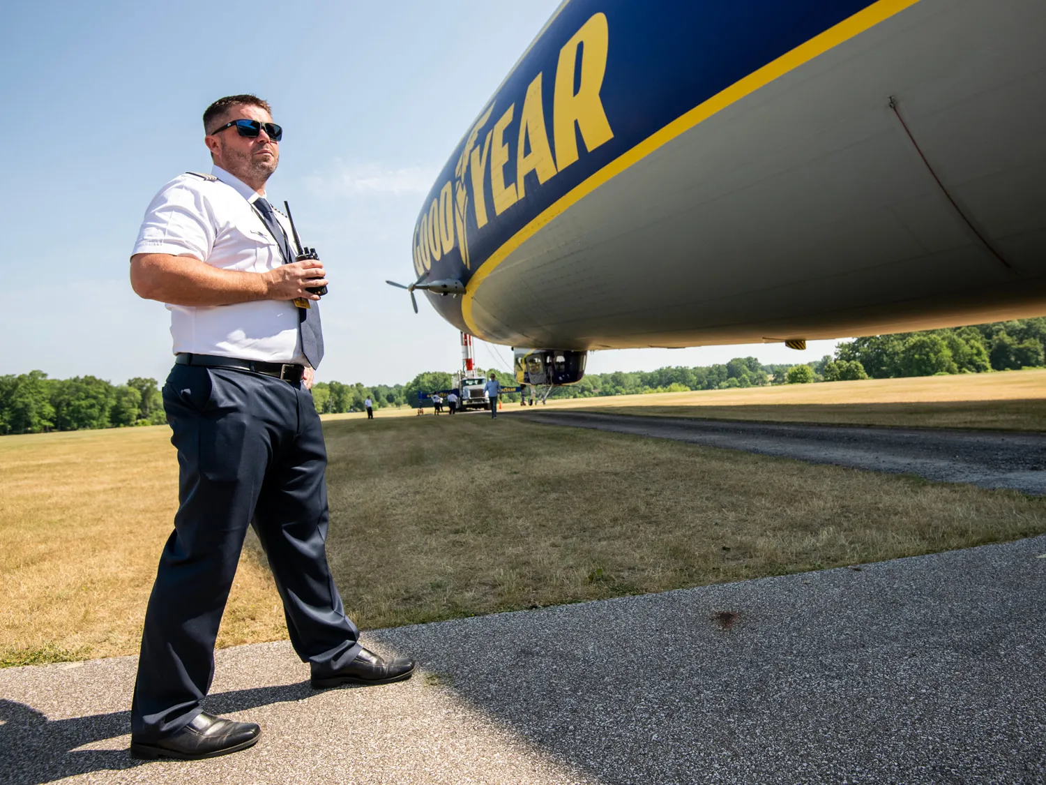 A man in a short-sleeved button down, dress pants, a tie and sunglasses holds a walkie talkie as the blimp is towed into position. 