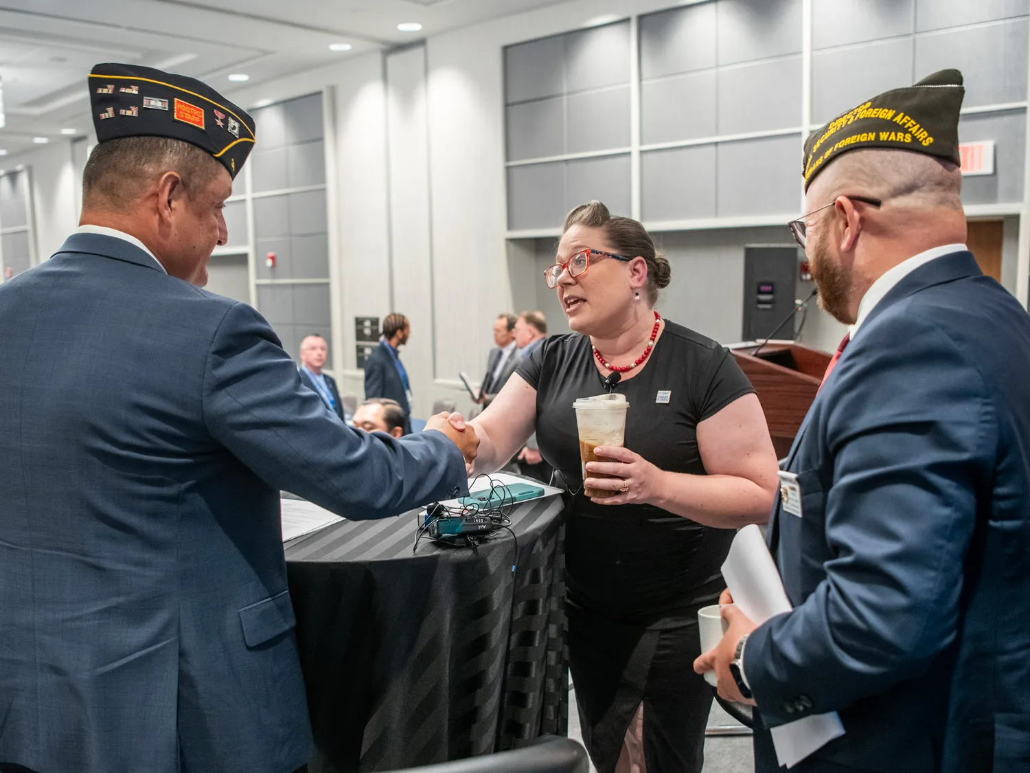 At a table on a small stage, Gretchen Klingler, a white woman with a style reminiscent of the 1940s, leans across a table to shake hands with a white man wearing an American Legion uniform cap and a suit. They’re making eye contact, and next to them smiling is a white man in a suit, this one with a VFW uniform cap. 