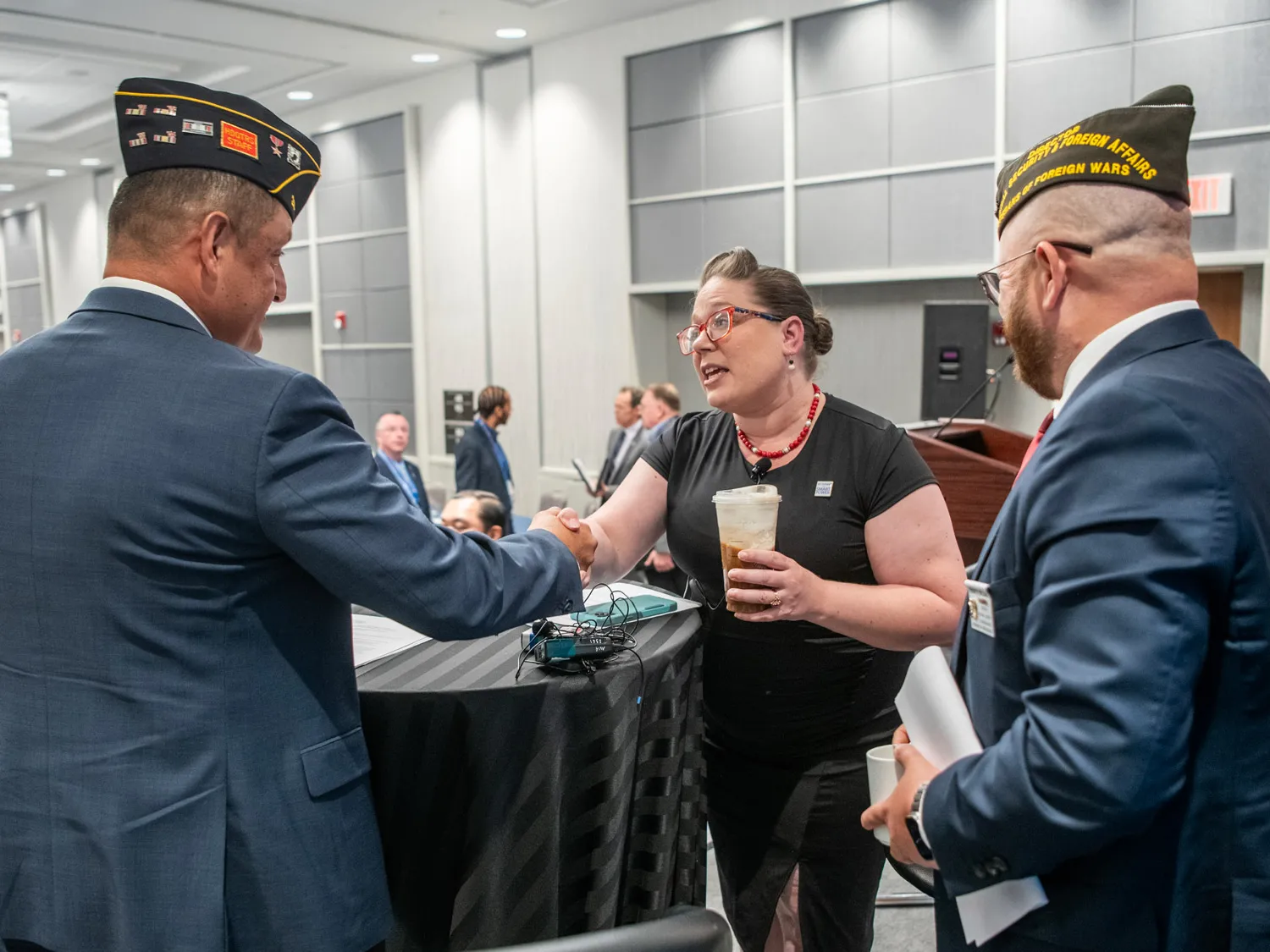 At a table on a small stage, Gretchen Klingler, a white woman with a style reminiscent of the 1940s, leans across a table to shake hands with a white man wearing an American Legion uniform cap and a suit. They’re making eye contact, and next to them smiling is a white man in a suit, this one with a VFW uniform cap. 