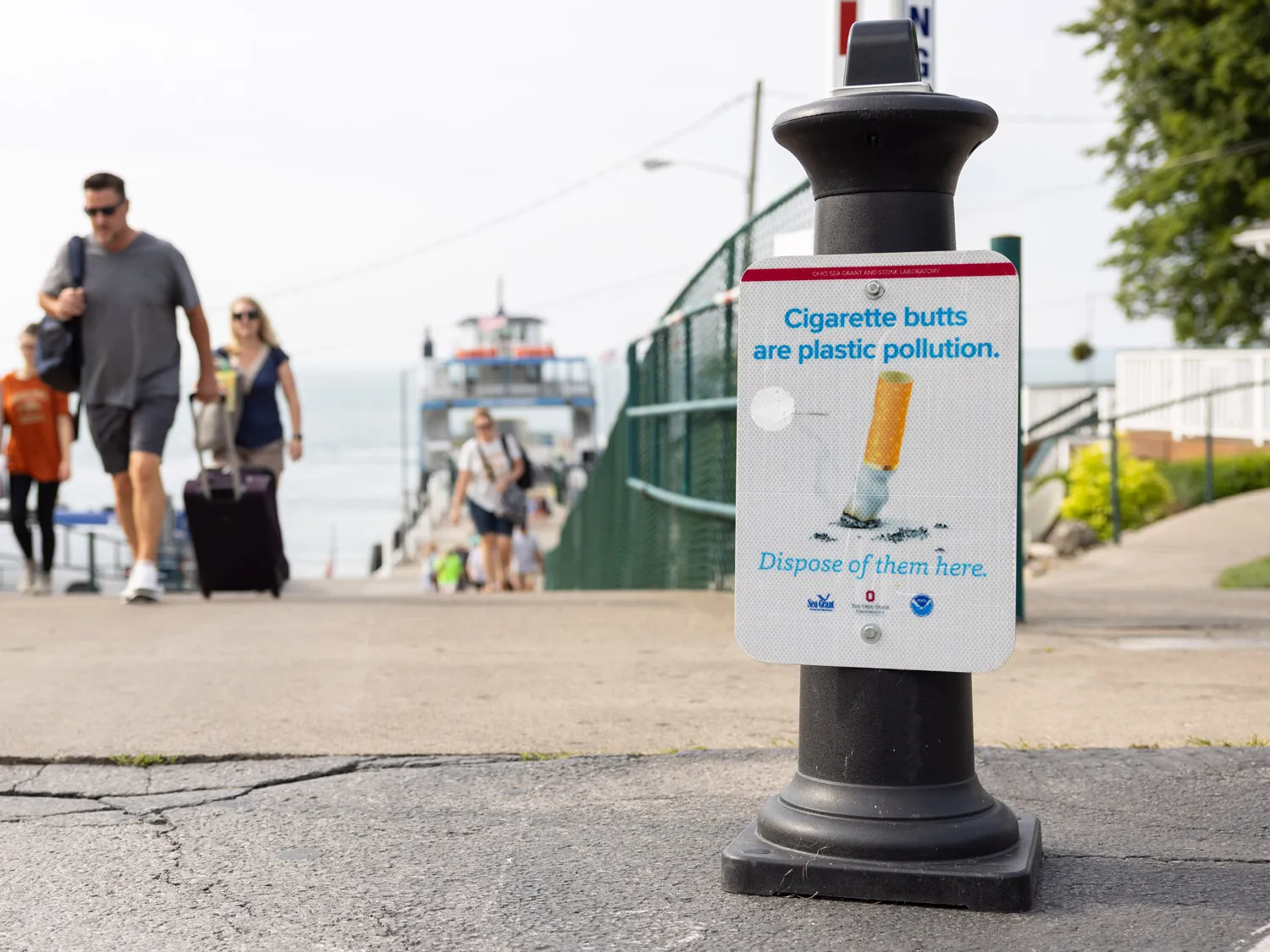 A sign says Cigarette butts are plastic pollution. Dispose of them here. The sign shows a cigarette butt being stamped out, and the trash receptacle it hangs on stands next to path where people walk onto the island from ferries. A handful of people are out-of-focus in the background.
