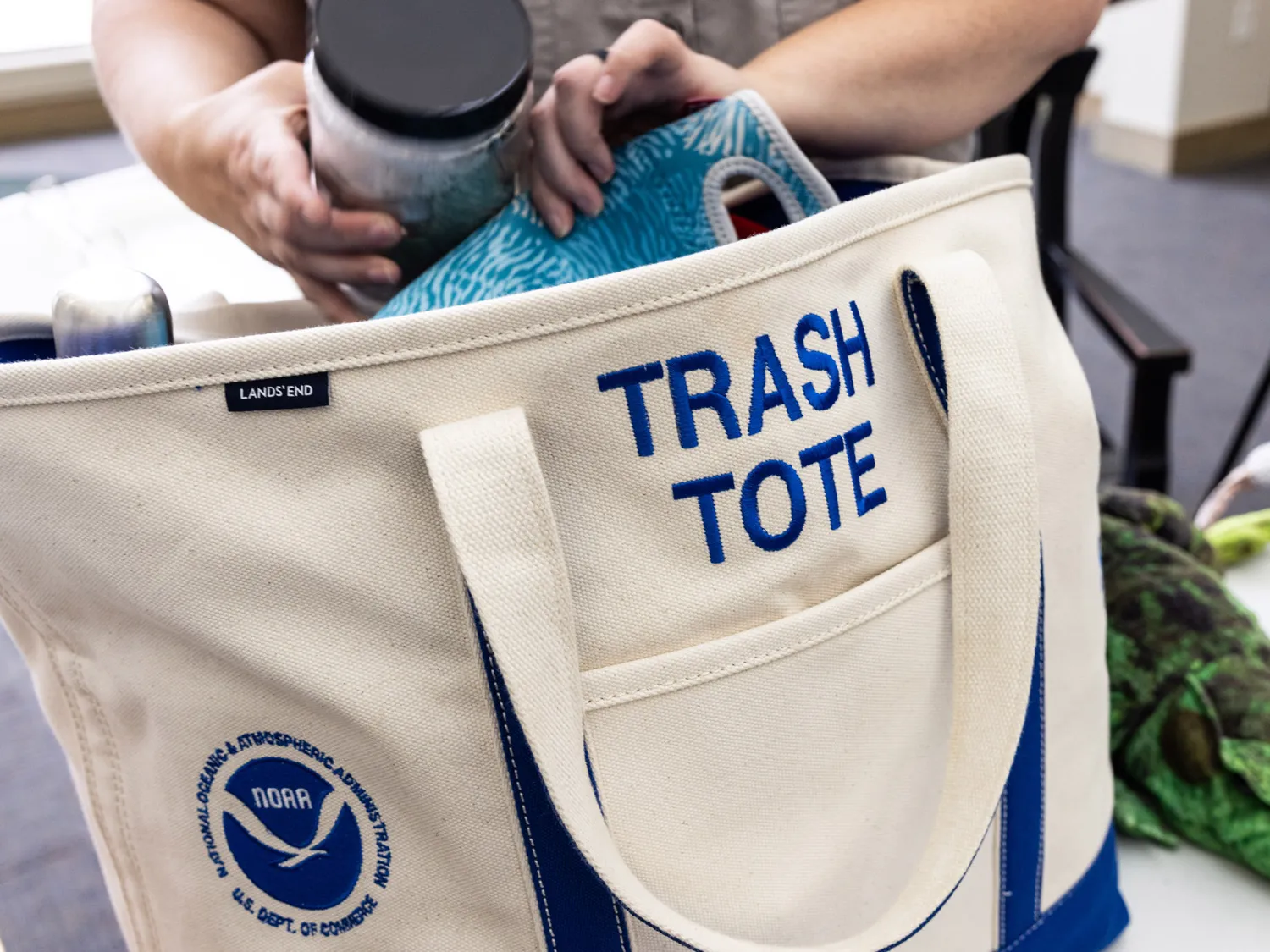 A large canvas bag has trash tote and a National Oceanic & Atmospheric Administration logo embroidered on the side. A person, whose hands are the only parts of them that can be seen, is picking up what seems like a plastic jar and reusable lunch bag, but it's unclear what is inside them.