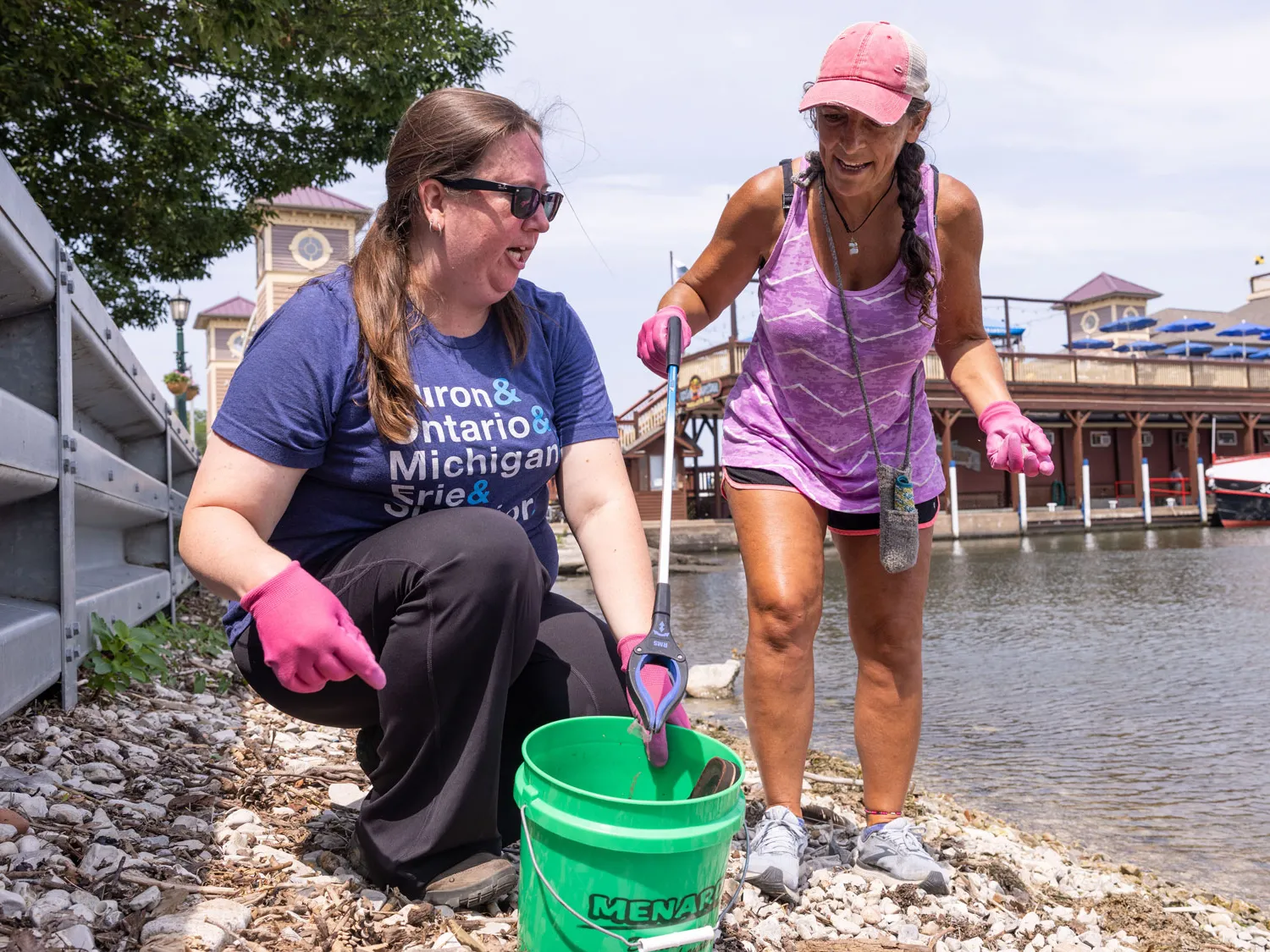 Between Lake Erie and a short metal wall, two women pick up trash on a rocky shoreline. They have a bucket to put trash in and wear gloves. One woman crouches down the other leans toward her with a pickup tool claw on a long handle. 