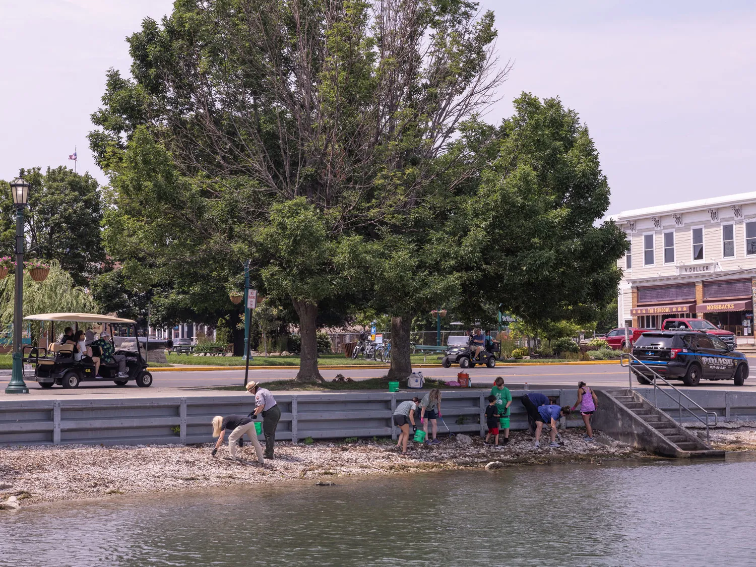 Taken from out in Lake Erie, a look into Put-In-Bay shows eight people on the rocky strip of beach, working next to a 4-foot wall. Above is the town, cars, a parking lot and giant trees. The people are picking up trash and dropping it in buckets.