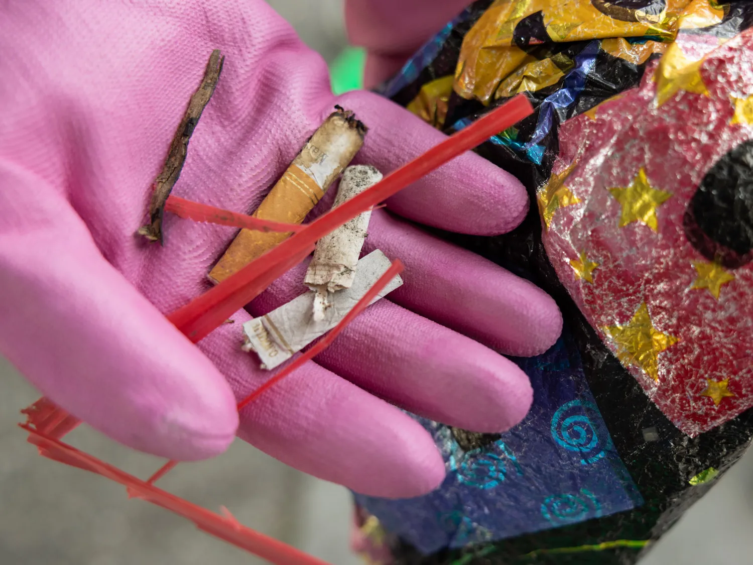 This photo shows someone's gloved hand holding trash-old straws, cigarette butts. In the background is a deflated mylar balloon, more trash picked up from the Put-In-Bay beach..
