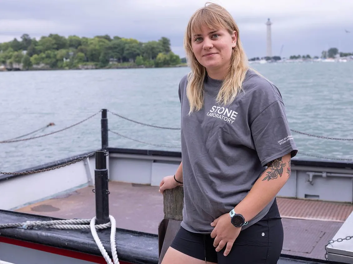A young blond woman stands in front of a boat and Lake Erie. She has a slight smile, a Stone Lab T-shirt and tattooed flowers on her left arm.