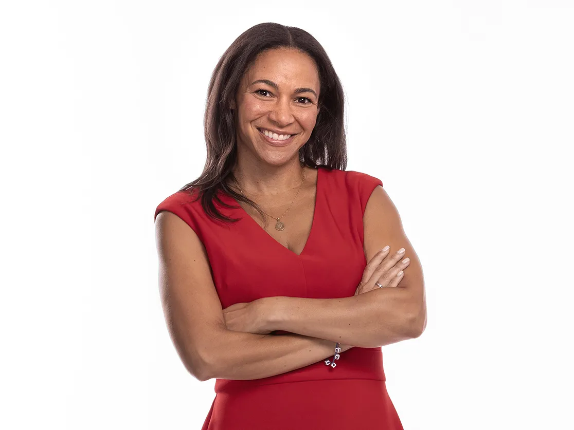 A woman wearing a scarlet dress smiles in a friendly, confident way as she poses in her high heels with her arms loosely crossed.