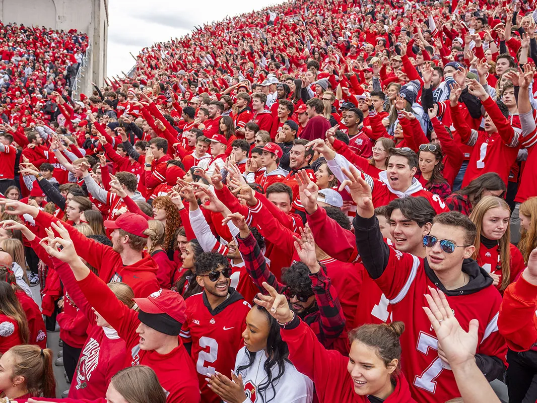 A sea of students in the stands at a football game wear Ohio State jerseys as they laugh and reach out while cheering on their team.