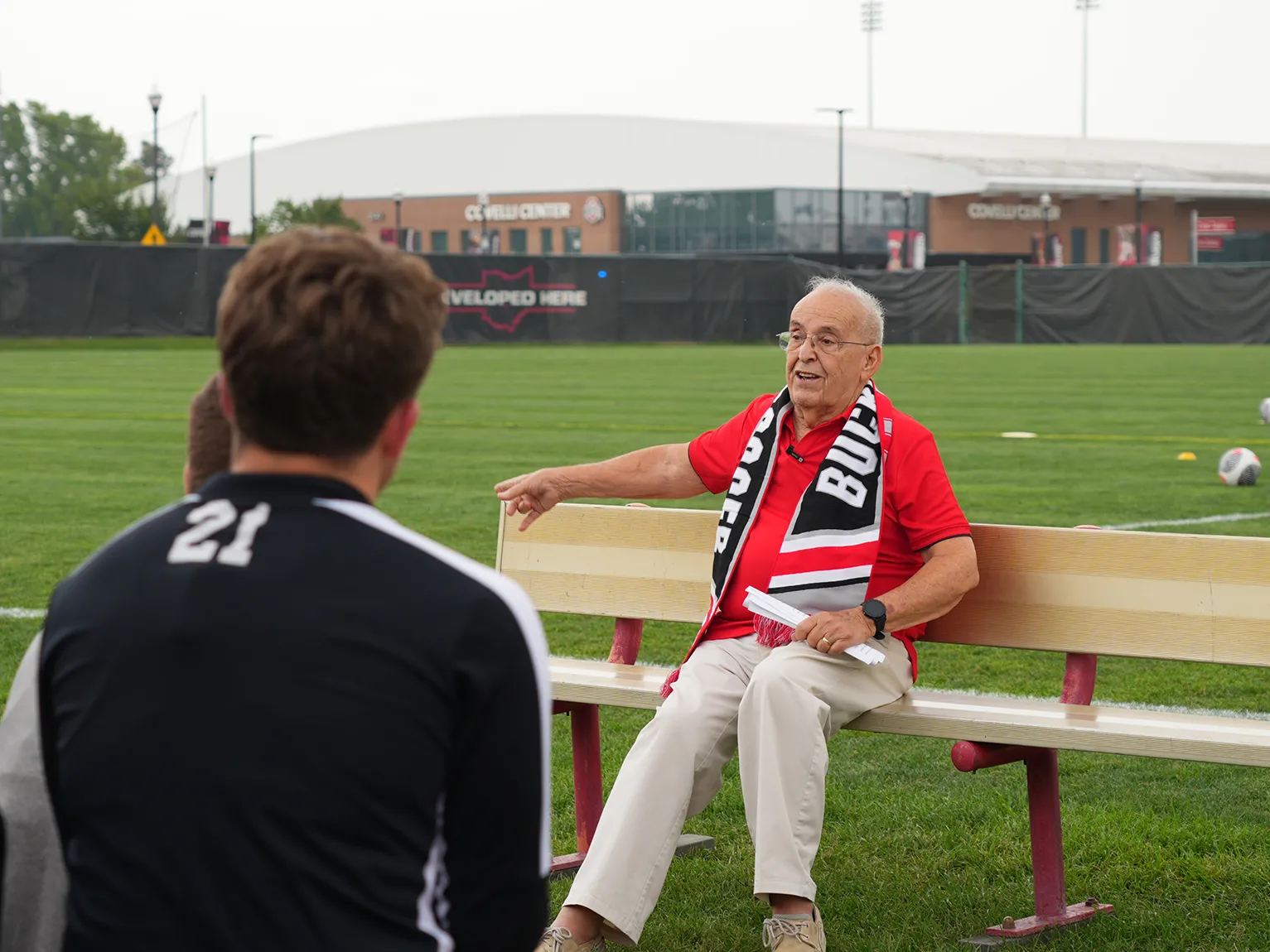 An older man happily chats, while wearing a scarlet shirt and long Buckeyes scarf, lounging on a bench along a soccer field at the Schumaker Complex. Behind him, the grass looks vibrant and soccer balls and field markings can be seen. Facing him are members of the men’s soccer team listening as he talks.