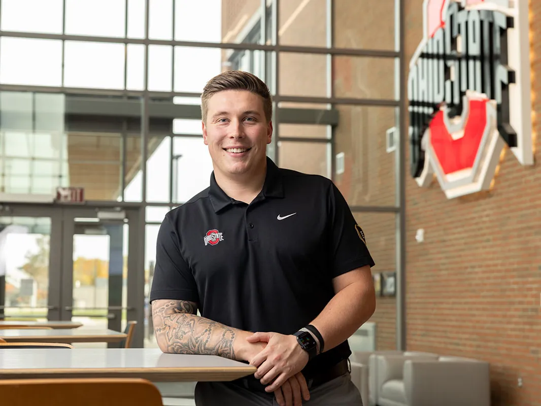 A young man wearing an Ohio State polo short smiles as he poses for this photo, leaning on a waist-height table in a building foyer where a large Ohio State logo hangs on the brick wall. He has neatly styled hair and tattoos on his right arm.