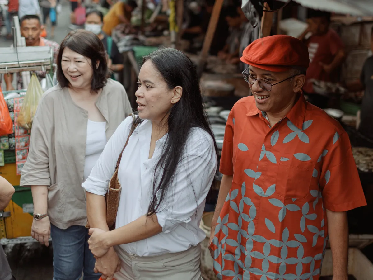 On a bustling street lined with stalls, three people take in an off-screen option. The man and two women smile as one of them comments on what it is they’re looking at.