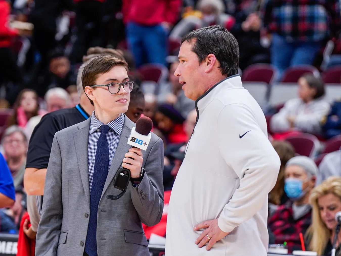 Standing on the sidelines of a basketball court, a young man in a suit and glasses listens intently and holds a microphone for the man he’s interviewing. That man, as adult in gym clothes, talk with hands on hips. Behind them, the stands are full with spectators wearing Ohio State colors.