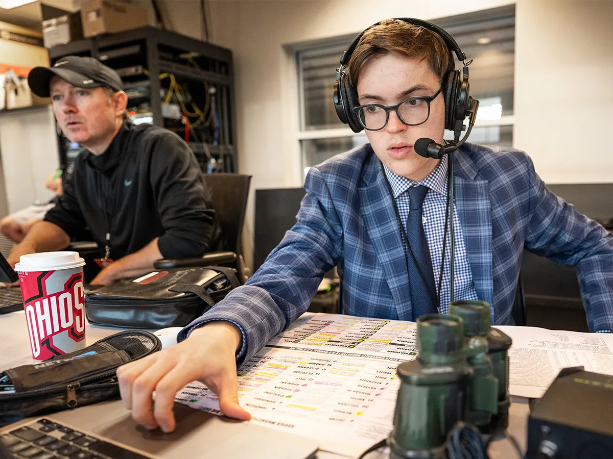 Wearing headphones with a mic, a checked sport coat and tie, Tyler Danburg focuses on game action off screen as he broadcasts a game from a the press box in a stadium.