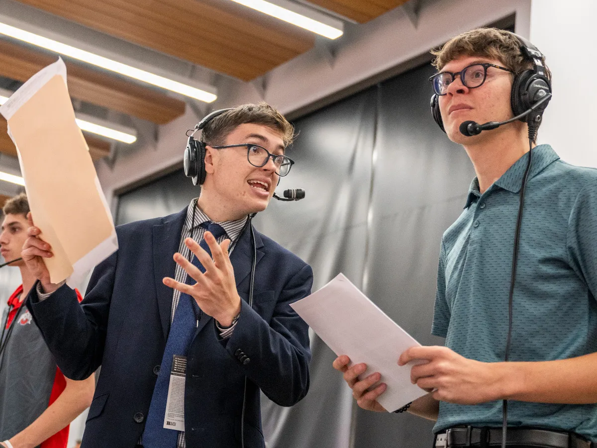 Tyler excitedly talks to a fellow game-caller. They both hold papers, wear headsets and intensely focus on what they're doing.