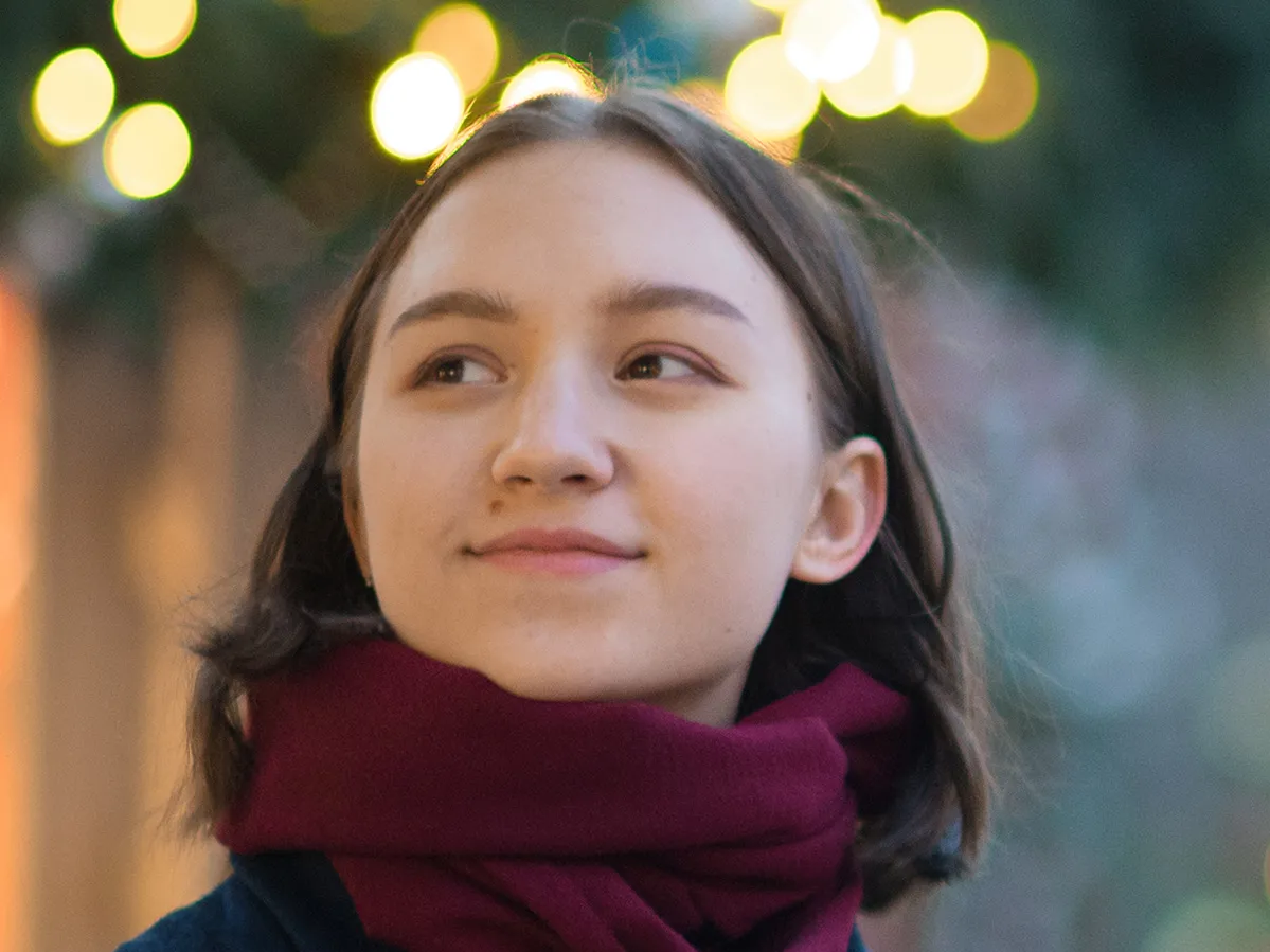 A young woman with a tightly bundled scarf and very slight smile stands among outdoor holiday lighting
