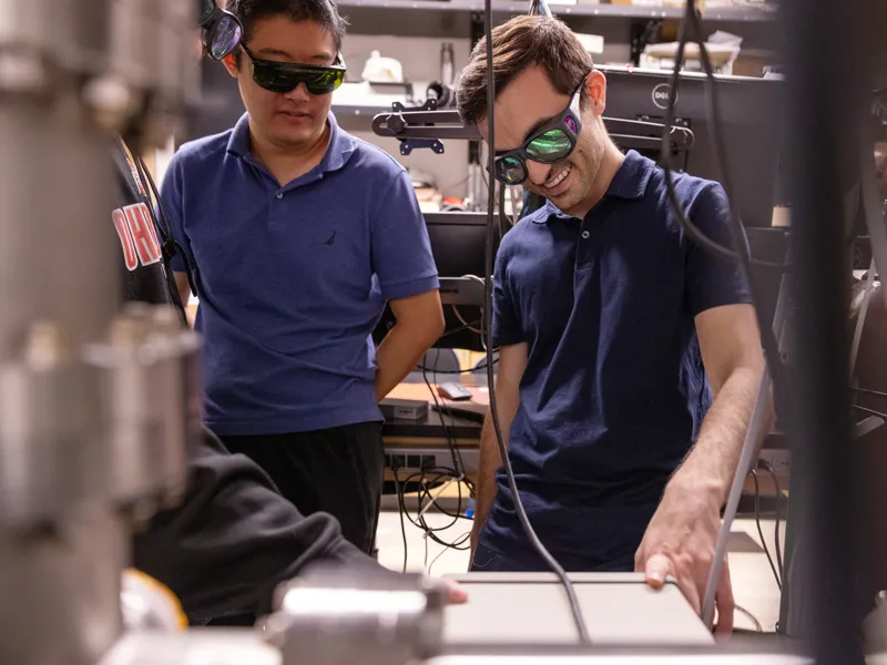 Two young men wearing blue shirts chat as they look at part of a machine that is mostly out of focus. They’re lab partners and both wear safety goggles.