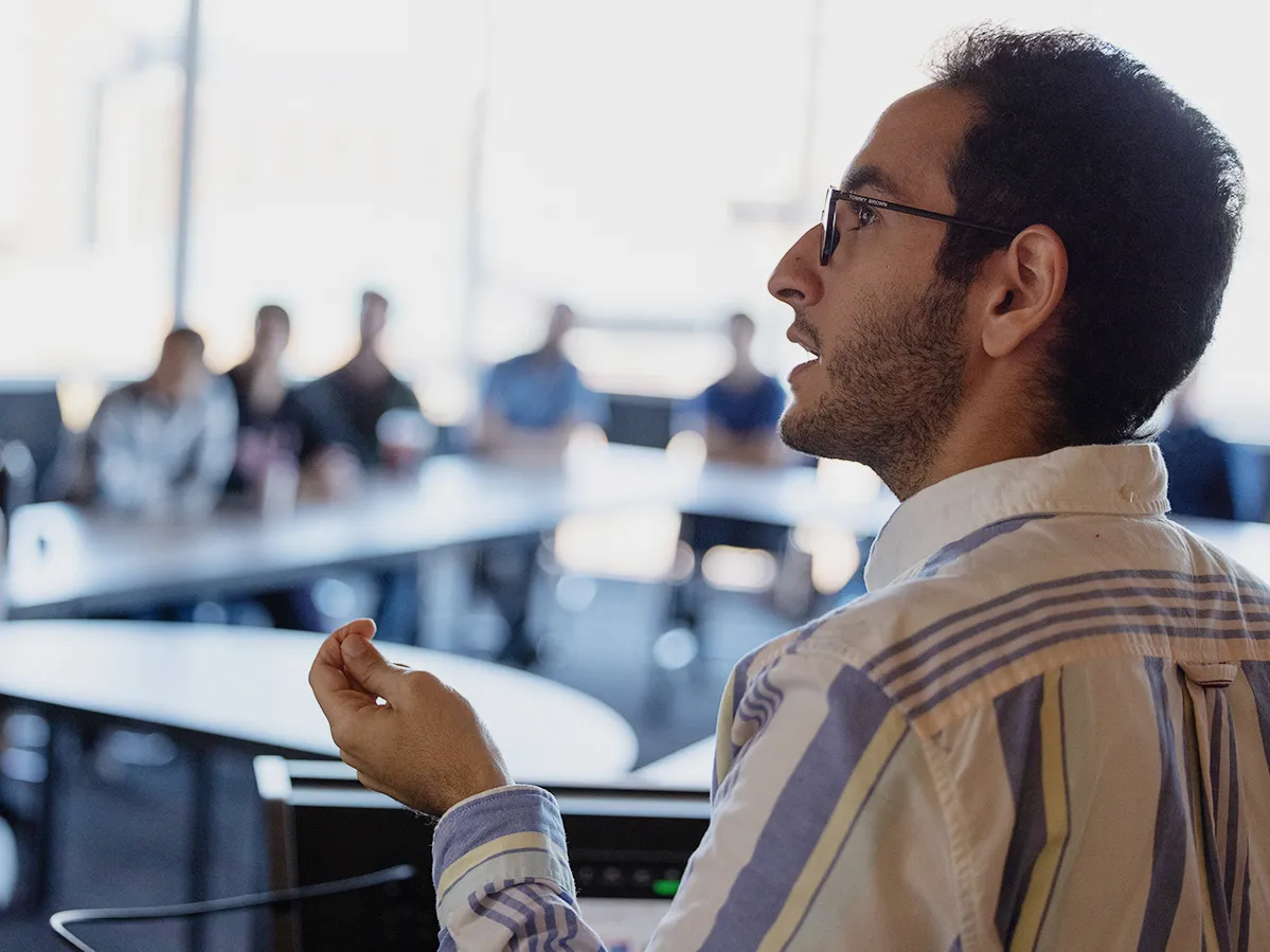 Abdallah AlShafey looks toward his slide presentation on a screen as he explains a point about the research he’s summarizing. Out of focus in the background, fellow students sit at conference tables listening attentively.