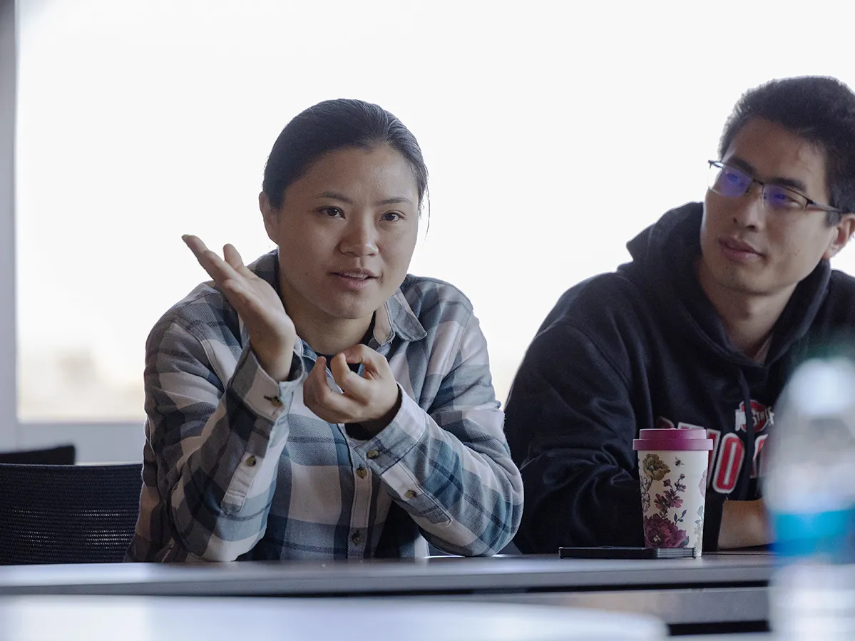 Post Doc Sha Li, who goes by Lisa, gestures as she asks a question during a meeting. She wears a plaid shirt and has her hair back in a ponytail while looking directly at the speaker with a slight smile as she talks. Nest to her is another Chinese student who watches her contemplatively.