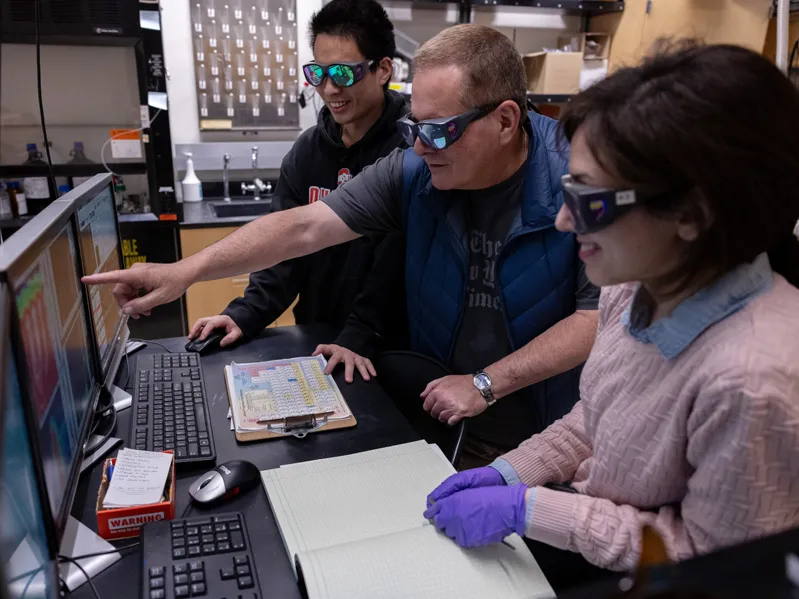 Three people—a professor and two of his students—look at a screen displaying colors and graphs that reflect how electrons moved in their experiments.