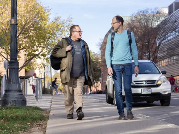 Two men walk up a sidewalk while leaving the physics building. One is shorter and carries a backpack over one shoulder, this is Lou. The other is taller and has his backpack on both shoulders. The day looks sunny but they’re wearing jackets and sweaters.