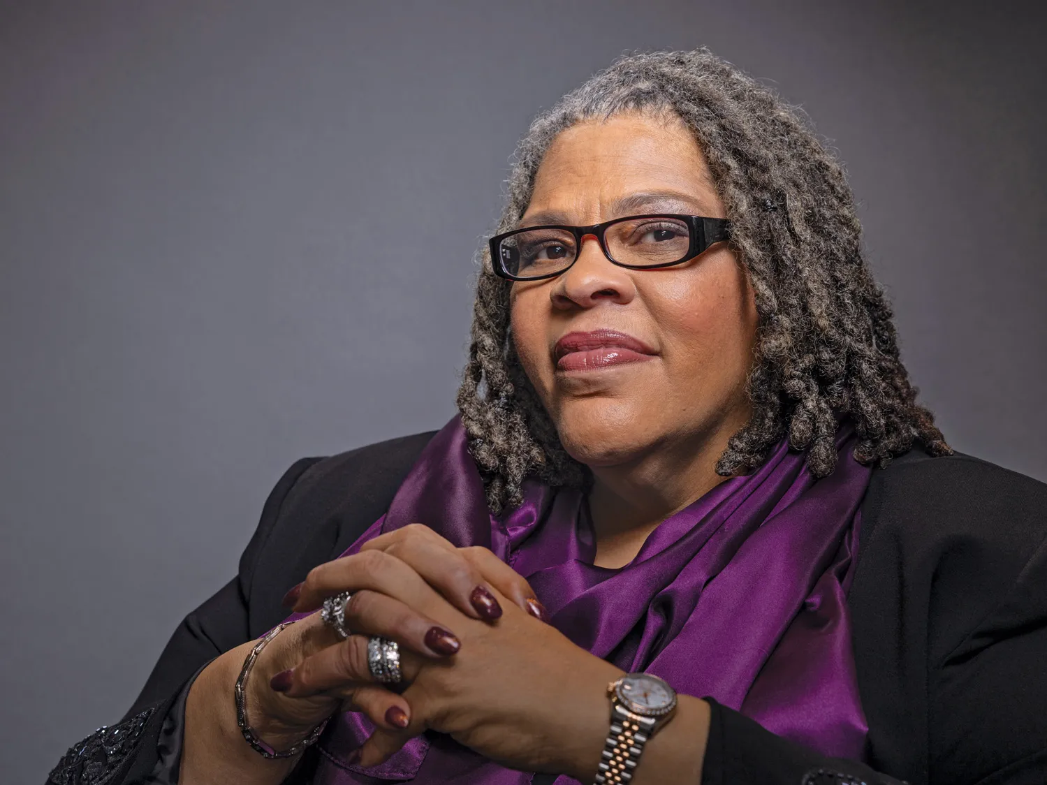 Patrice Palmer seems proud, thoughtful and friendly but direct as she poses for this photo with a slight smile that nevertheless lights up her face. She seems to be looking the viewer in the eye. She is an older Black woman with shoulder-length hair and wears rectangular glasses, a business suit, a silk scarf and silver rings and watch. 