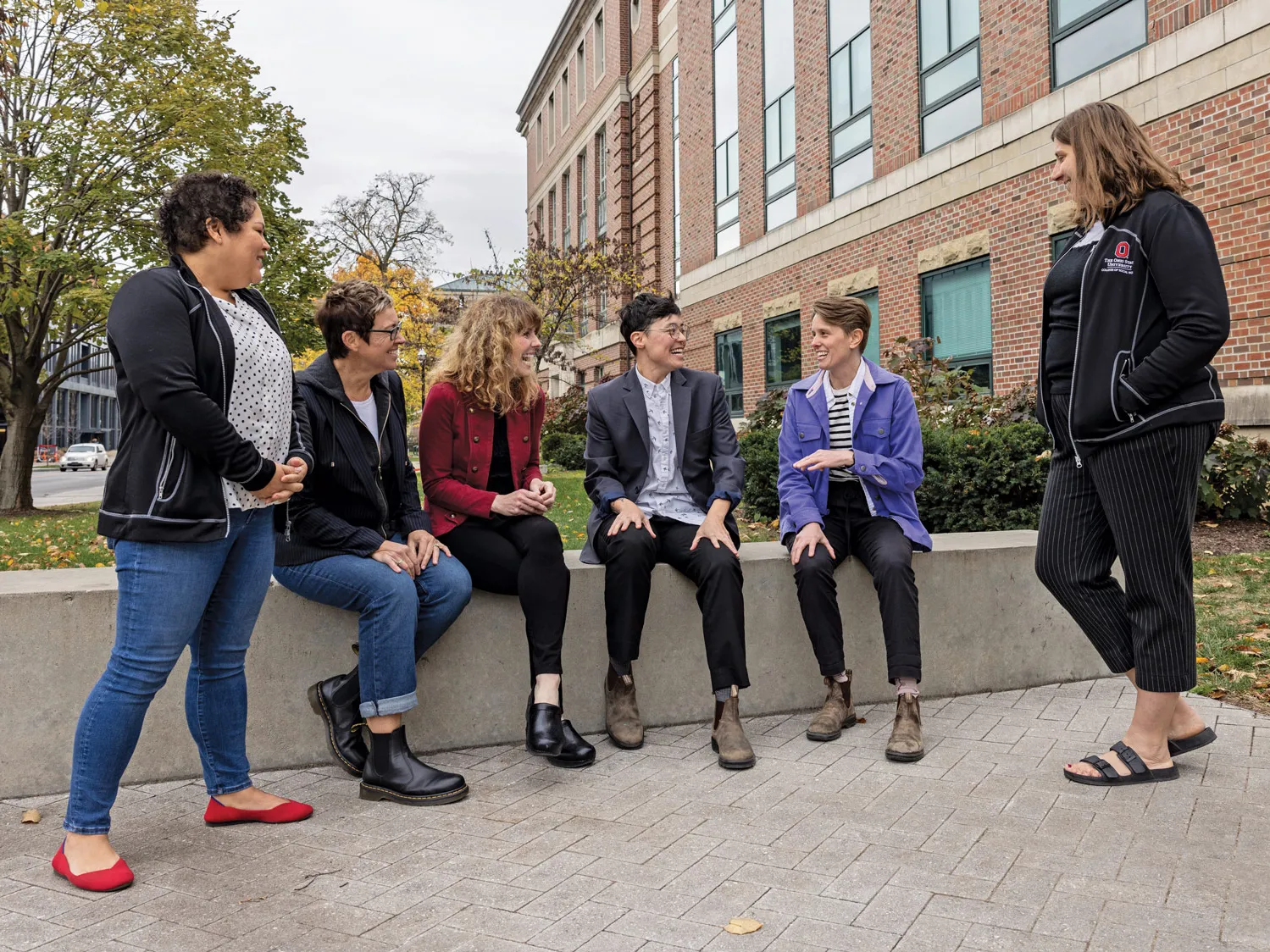 Outside of the brick Stillman Hall, a group gathers on a small paved plaza. Some sit on a low wall and some stand as they happily chat on what seems to be a chilly fall day, judging by their jackets and leaf colors on trees in the background. The people are a mix of ages and genders. 