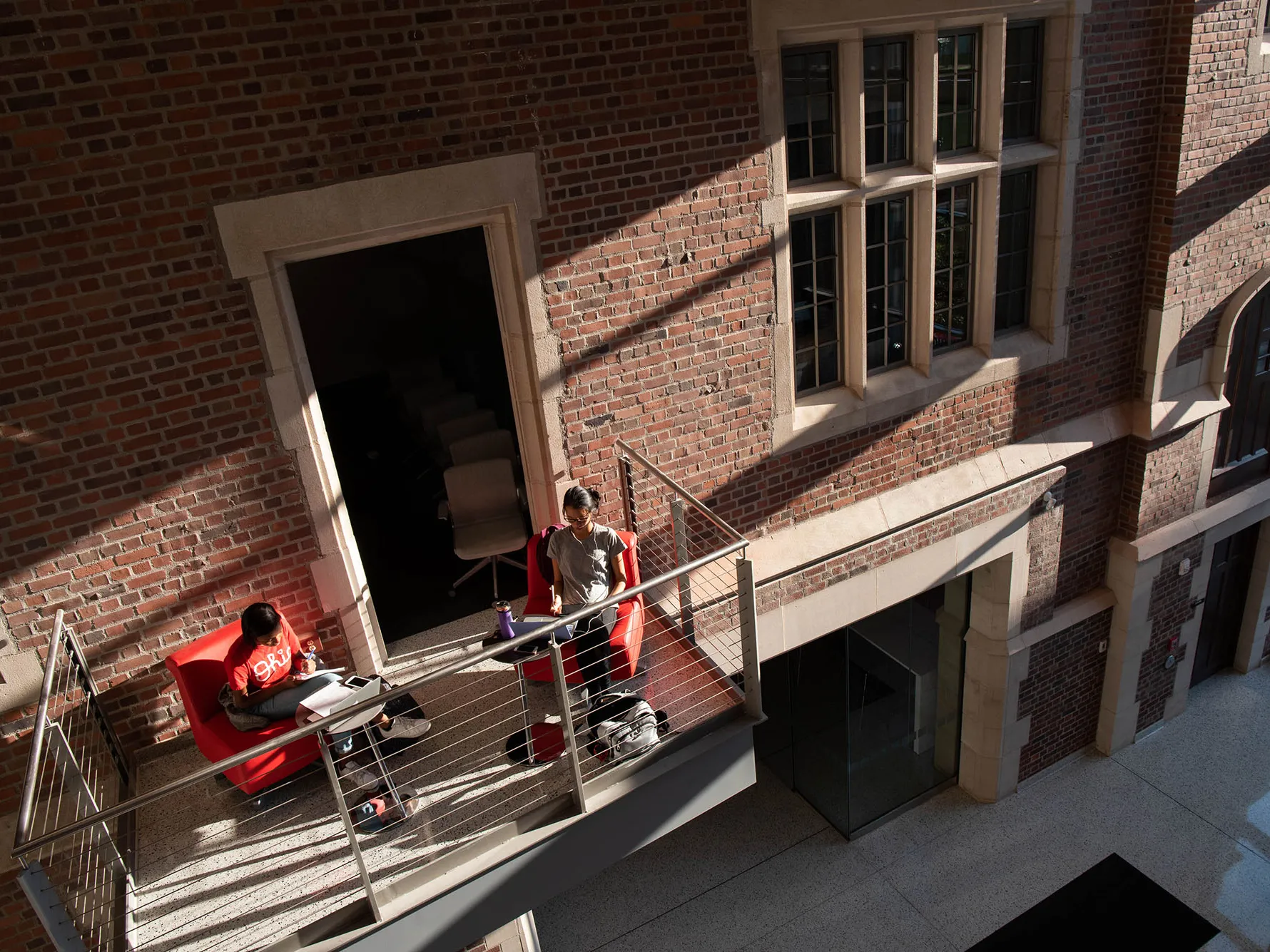 This photo shows Pomerene Hall on Ohio State’s campus. Inside, the lights are all on, so the windows glow—a geometric-pattern mural can be seen inside a wall of ground-to-roof windows. Students wearing backpacks pass by on the sidewalk in front.