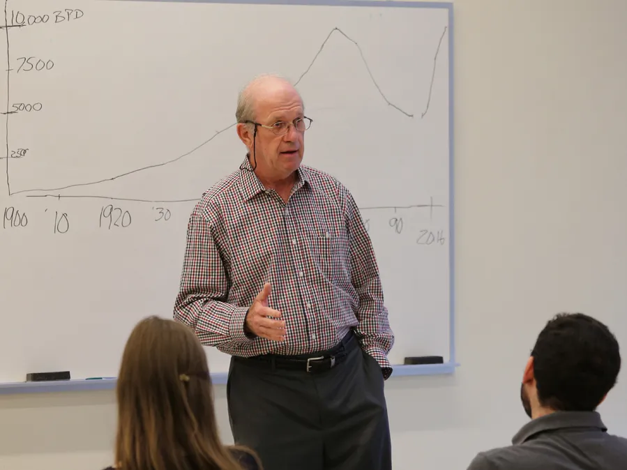 An older man wearing glasses and a checked button-down gestures while teaching a class in front of a white board.