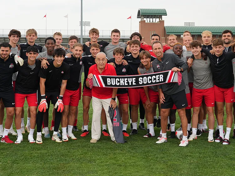 Paul Halpern, a 91-year-old man, poses with Ohio State&#039;s men&#039;s football team and coach on the soccer field at Schumaker Complex. They hold a banner that reads Buckeye soccer