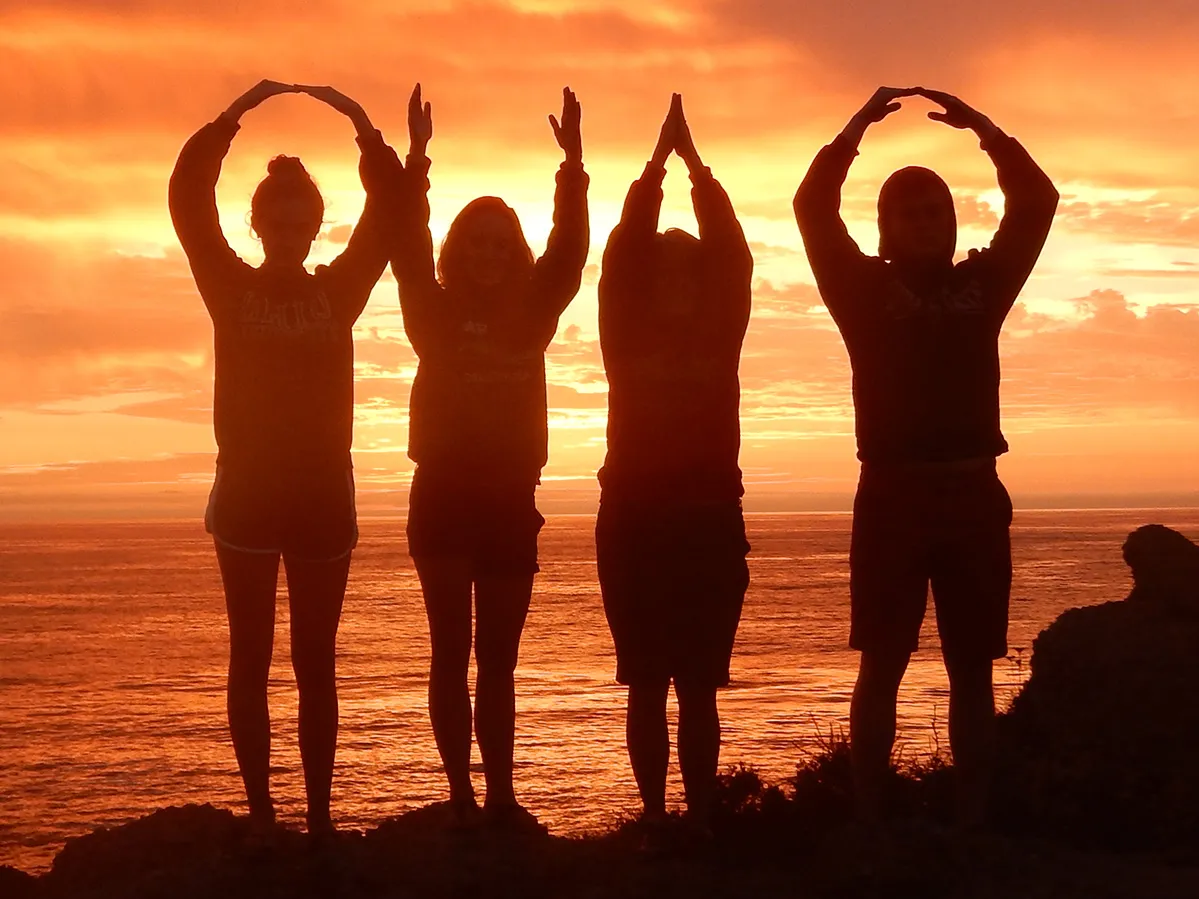 As the setting sun lights up the evening sky in shades of gold and orange, four people form an OHIO. Because of the backlighting, they appear solely as dark silhouettes.