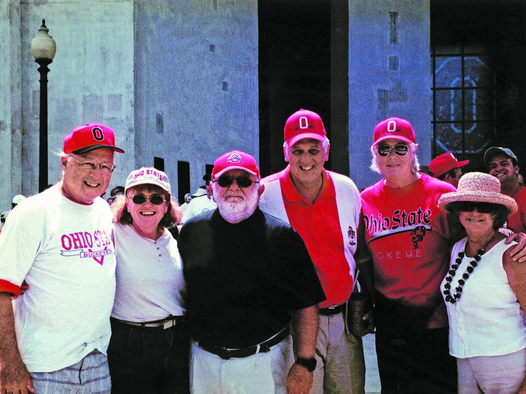 Three couples pose in front of the entrance to Ohio Stadium. They are all white, middle-aged people wearing Ohio State gear and baseball caps (though one lady has on a brimmed sunhat) who stand smiling with their arms around the shoulders of the people next to them.