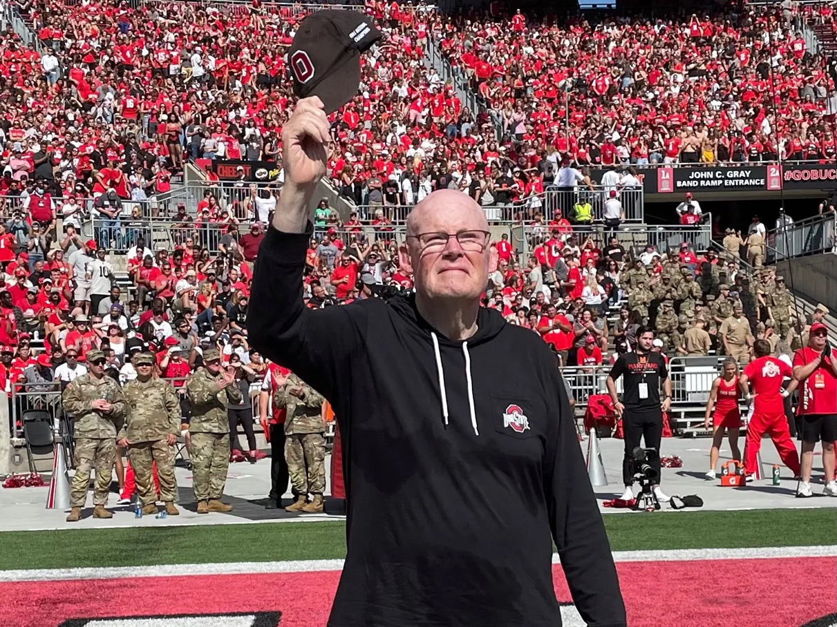 Standing on the field inside Ohio Stadium, Daniel Connor, an older white man dressed in matching pants and sweatshirt—with an Ohio State logo—waves his baseball cap in the air as he greets the crowd at a football game.