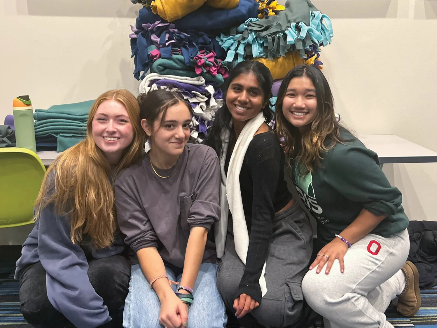 Crouching to pose in front of a table stacked high with fleece blankets are four female students smiling widely. The way they scooted close together speaks to the friends being very comfortable with one another. The blankets are double layered with wide fringe hand-knotted along each side.