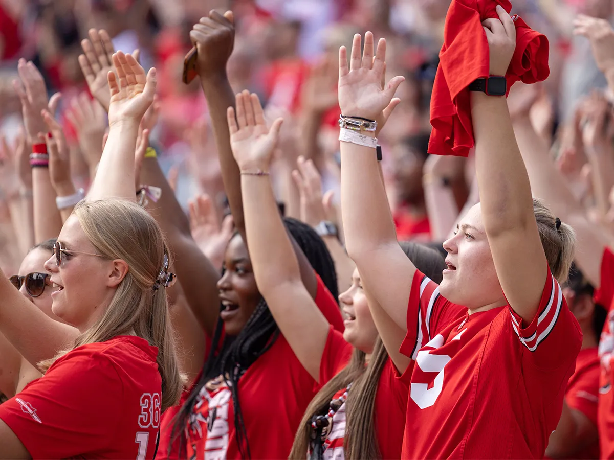 A group of fans among a stadium crowd raise their hands above their head and sing Carmen Ohio.