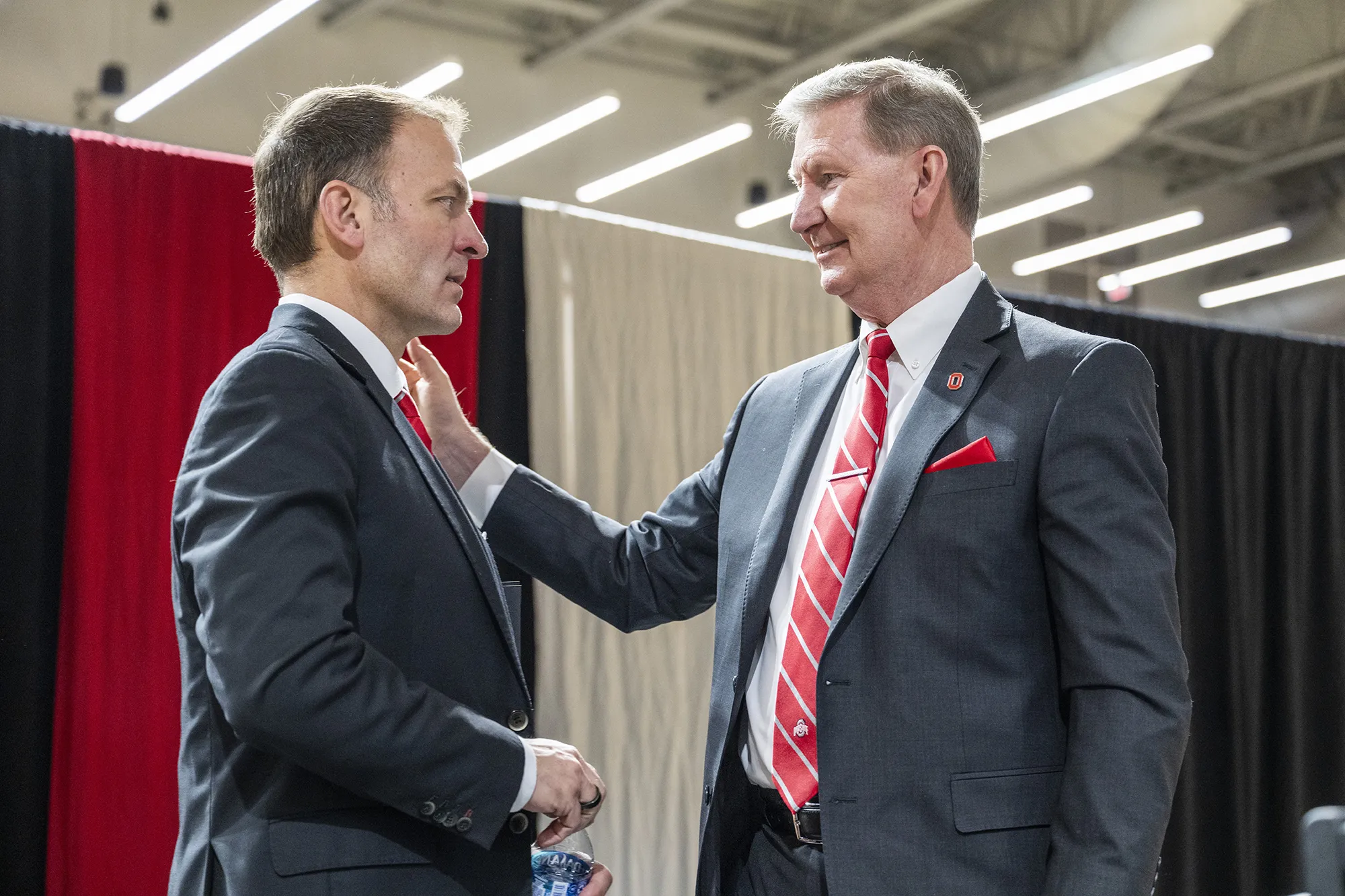 Ohio State President Ted Carter smiles and pats the shoulder of incoming athletic director Ross Bjork before a press conference to introduce him. Behind them are curtained panels. Both are white men in similarly colored suits and ties, but where President Carter seems relaxed, Bjork, in this moment the camera caught, seems as if he might be a bit nervous. 