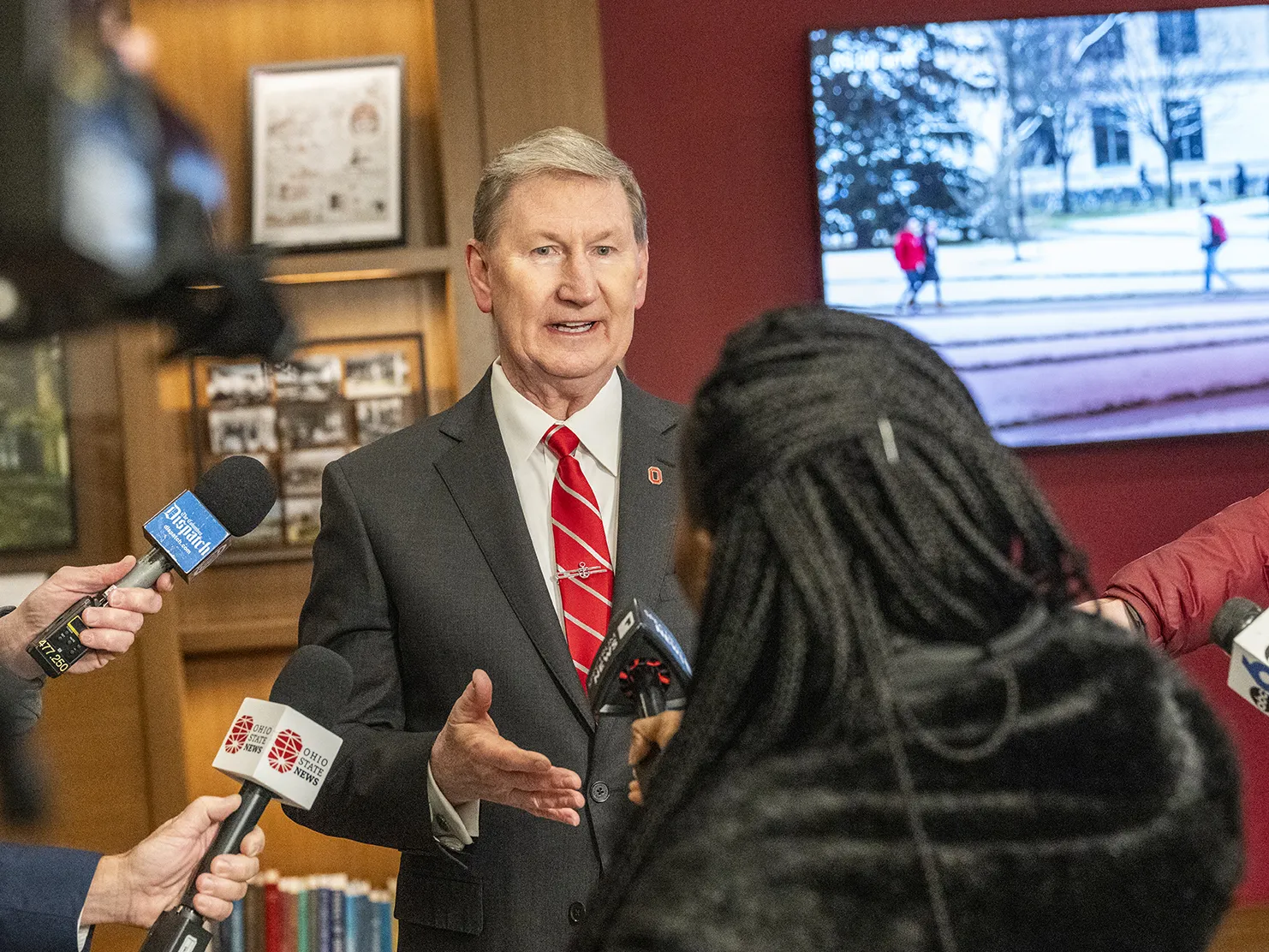 President Carter gestures as he answers questions from reporters circled around him. Most of the reporters don’t appear in the photo, but their outstretched arms holding microphones or cameras are aimed at him. The main reporter Carter is speaking to can be seen from behind. She has long hair in thin braids.