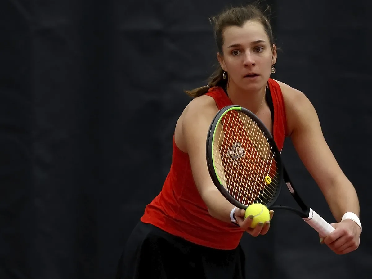 On the court, Irina Cantos Siemers, an Ohio State tennis player, holds a ball in her right hand and her racket in her left, getting ready to serve. She seems focused on her opponent’s side of the court as she prepares for action. 