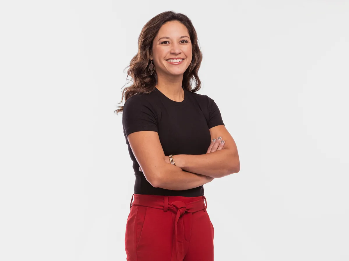 Stephanie Baxter, a white woman with long wavy hair, stands up straight, smiling with her arms crossed. She comes across as happy and confident as she poses in a fitted T-shirt, fashionable pants and heels.