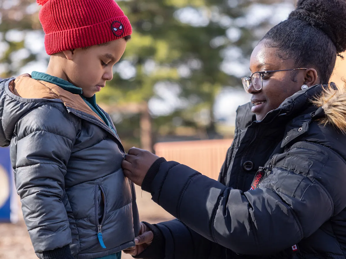 On an outdoor playground, a college student smiles as she crouches down to zip a child’s coat. She’s also wearing a heavy winter coat (along with glasses and her hair piled in a big bun on top of her head). The boy, wearing a stocking hat and gloves, watches her hands.