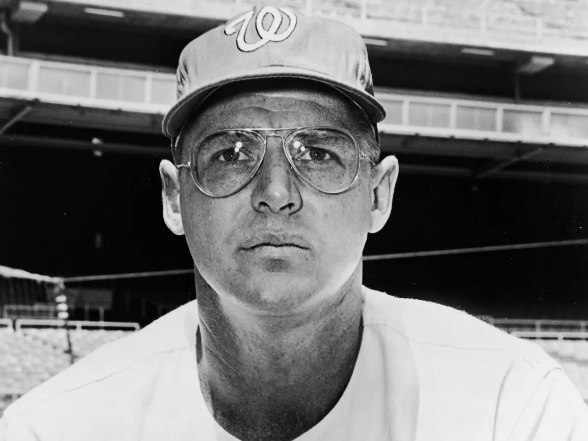 In an older photo, a white man leans toward the camera while wearing his baseball uniform, glasses and a baseball cap. He looks focused and serious.