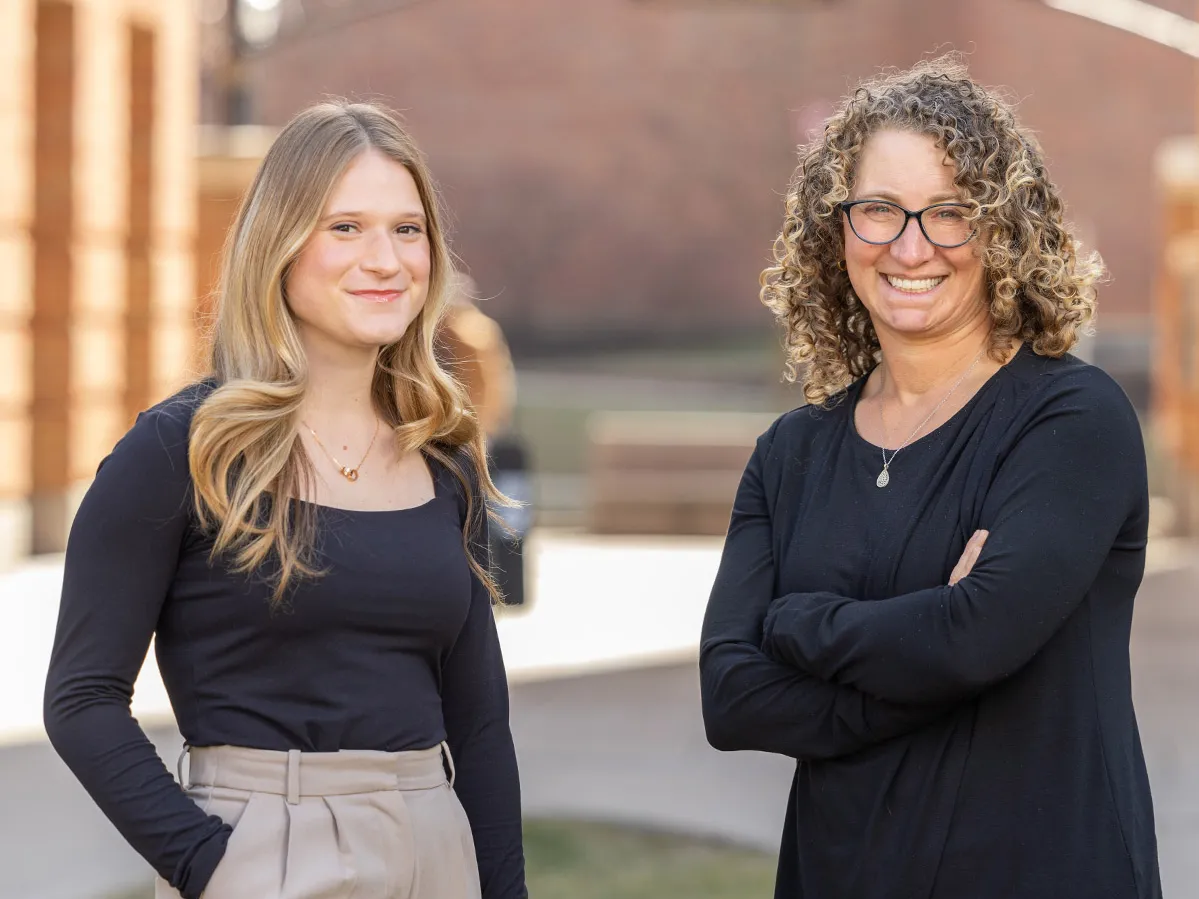 Two women stand in the courtyard of Ohio State’s Fisher College of Business. Both wear dark tops. The college student, on the left, has long blond hair and stands with one hand tucked in her pocket. The teacher, on the left, has curly shoulder length hair and stands with her arms crossed. Both smile as they pose.