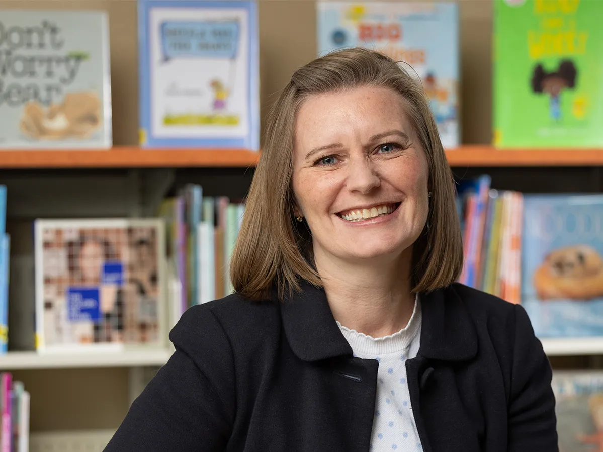 Shayne Piasta poses in a reading room surrounded by children’s books. Coming off as friendly and direct, she sits at a small table with her hands folded. She has a wide smile that crinkles the skin around her eyes and shoulder length hair.