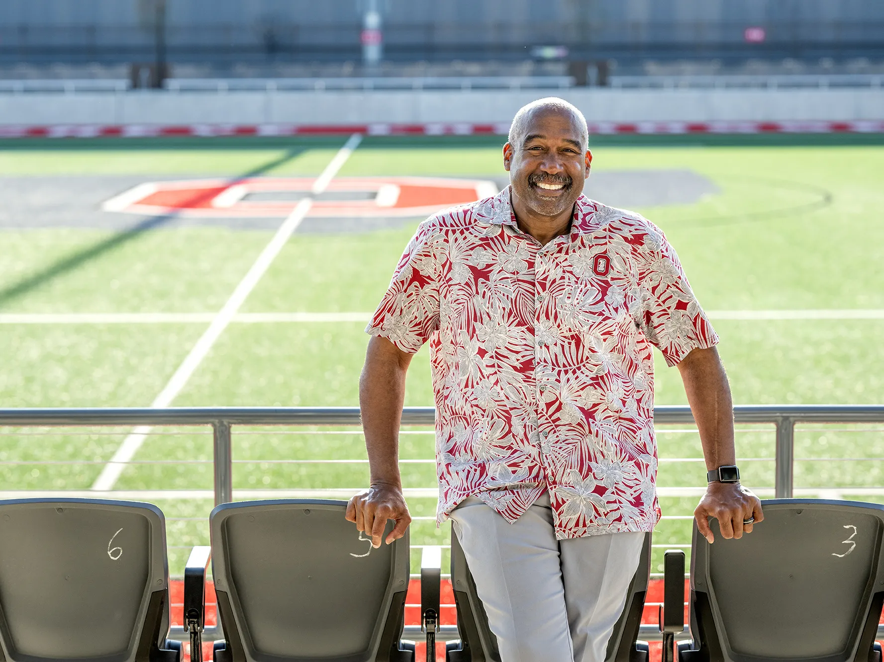 Wearing an Ohio State-colored tropical shirt—that looks perfect for retirement or vacation—Gene Smith grins as he leans on some front-row seats at Ohio Stadium.