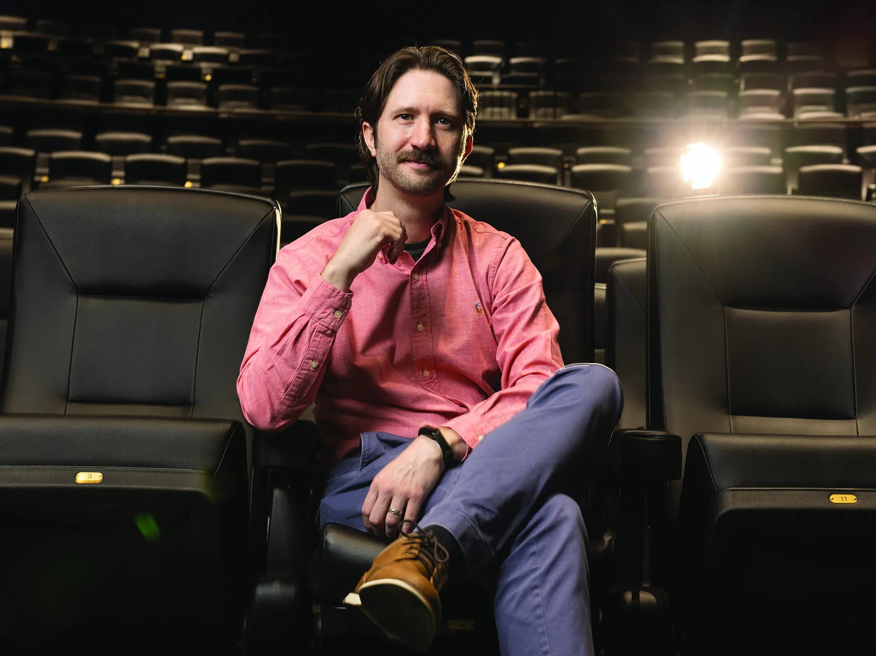 Sitting in a theater with leather seats, Matthew Grizzard, a bearded white man wearing khakis and a button down, sits relaxed with one leg crossed over the other and his hands seeming as if he’s focusing as he listens. He’s smiling with his eyes more than his mouth.