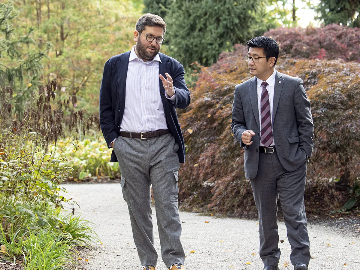 Walking on a gravel path through an area with lush plants and trees, Jeffrey Schottenstein, a bearded white man wearing a cardigan, button-up and glasses, gestures as he speaks with Dr. K Luan Phan, a man of Asian descent wearing a suit and tie. He’s listening intently and the men’s heads are canted toward each other as they look forward.