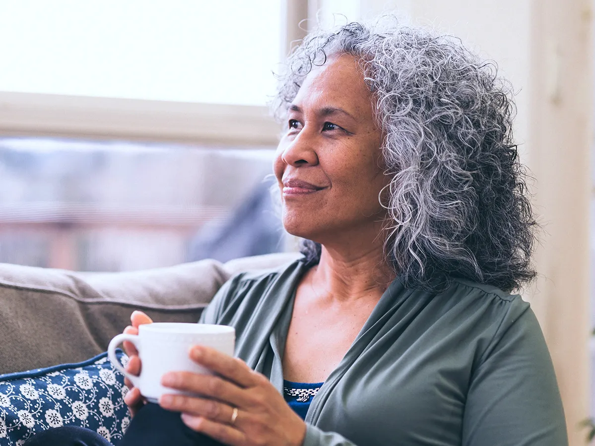 A woman sits with knees to her body near a window and holding a hot beverage. She appears contemplative as she looks into the distance